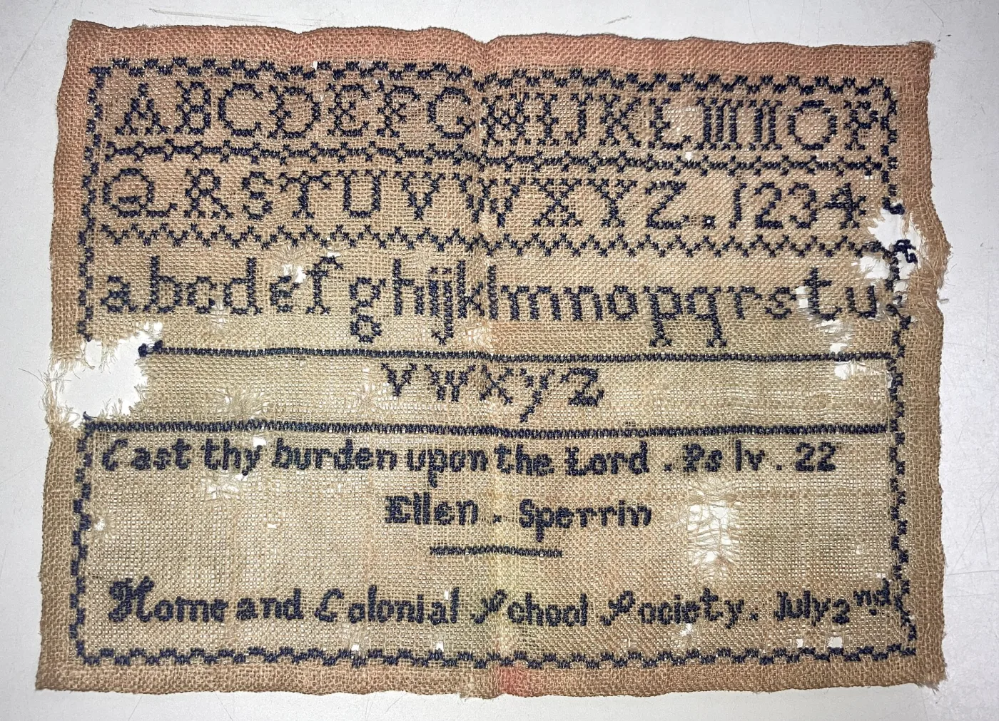 Small unmounted sampler on coarse tan linen worked in dark blue thread, with two alphabets, numerals, a scripture verse, a signature and a school name, and losses at the edges