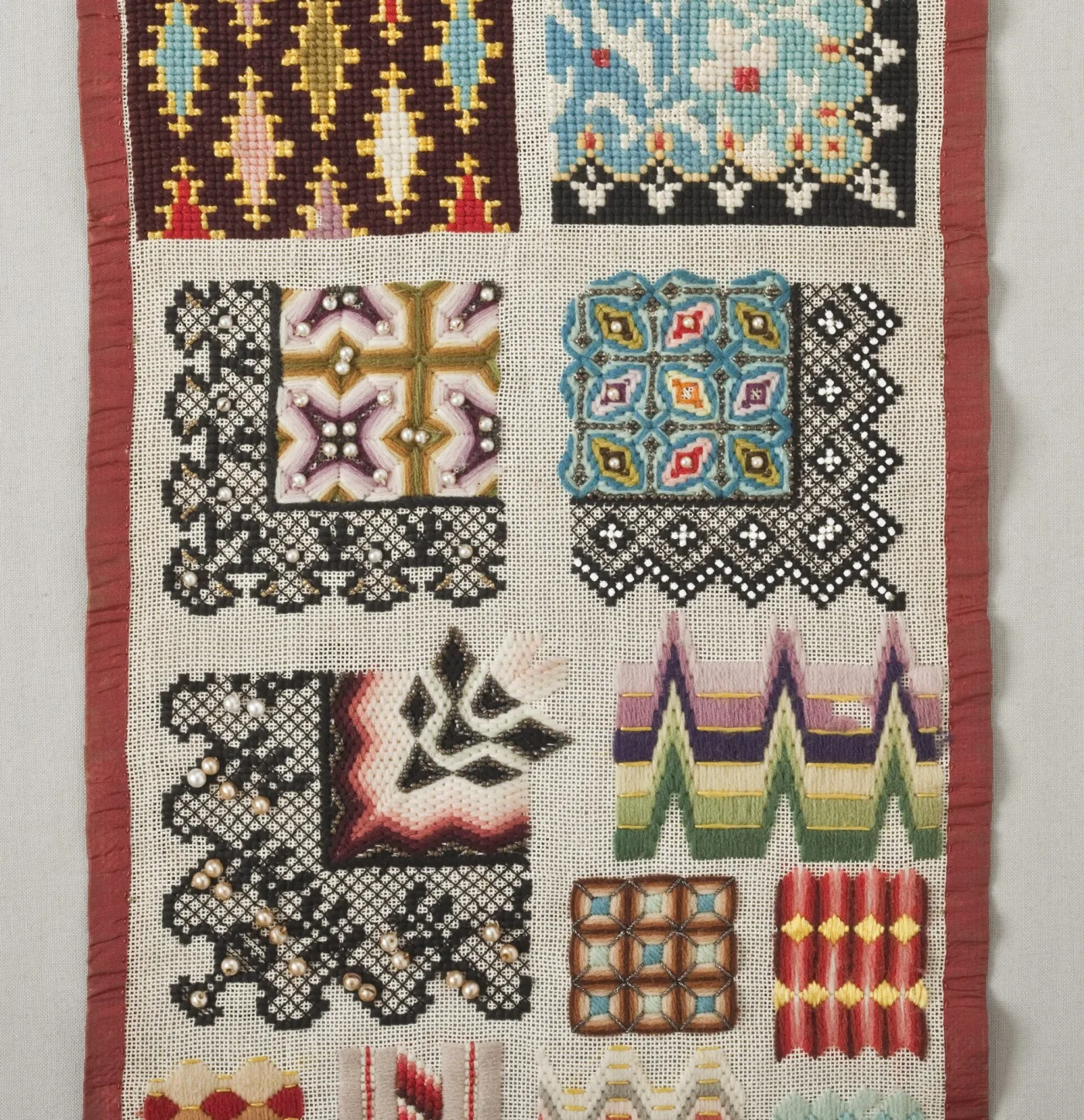 Detail of a narrow sampler densely worked in coloured wools on canvas, with squares of geometric pattern, black lace-like open stitching, and rows of seed pearls and glass beads