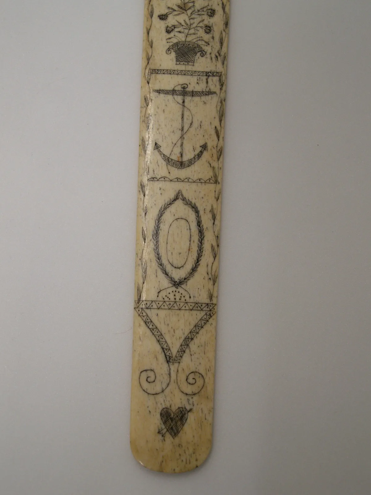 Upper end of a whalebone busk standing vertically, engraved with a foliate crest above a panelled anchor and an oval reserve, framed by ruled borders