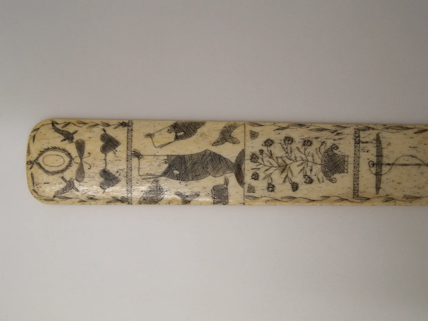 Long flat strip of pale whalebone engraved along its length with panels containing a fish, a heart, foliage and geometric borders, the edges slightly waisted