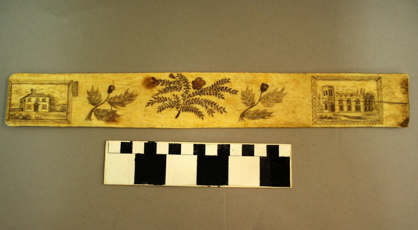 Yellowed whalebone busk laid horizontally beside a black and white scale bar, engraved with a building at one end, sprays of fern and leaves along its length and a second building at the other