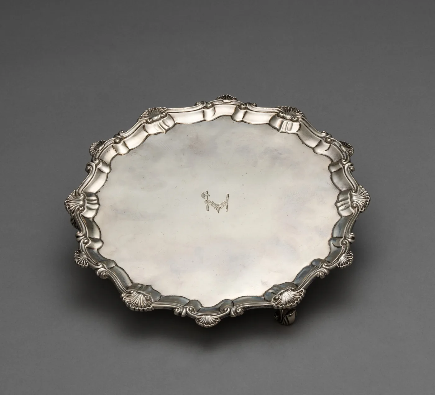 Circular silver salver with a cast rim of alternating shells and scrolls, an engraved armorial device at the centre, and a small scrolled foot visible beneath one edge