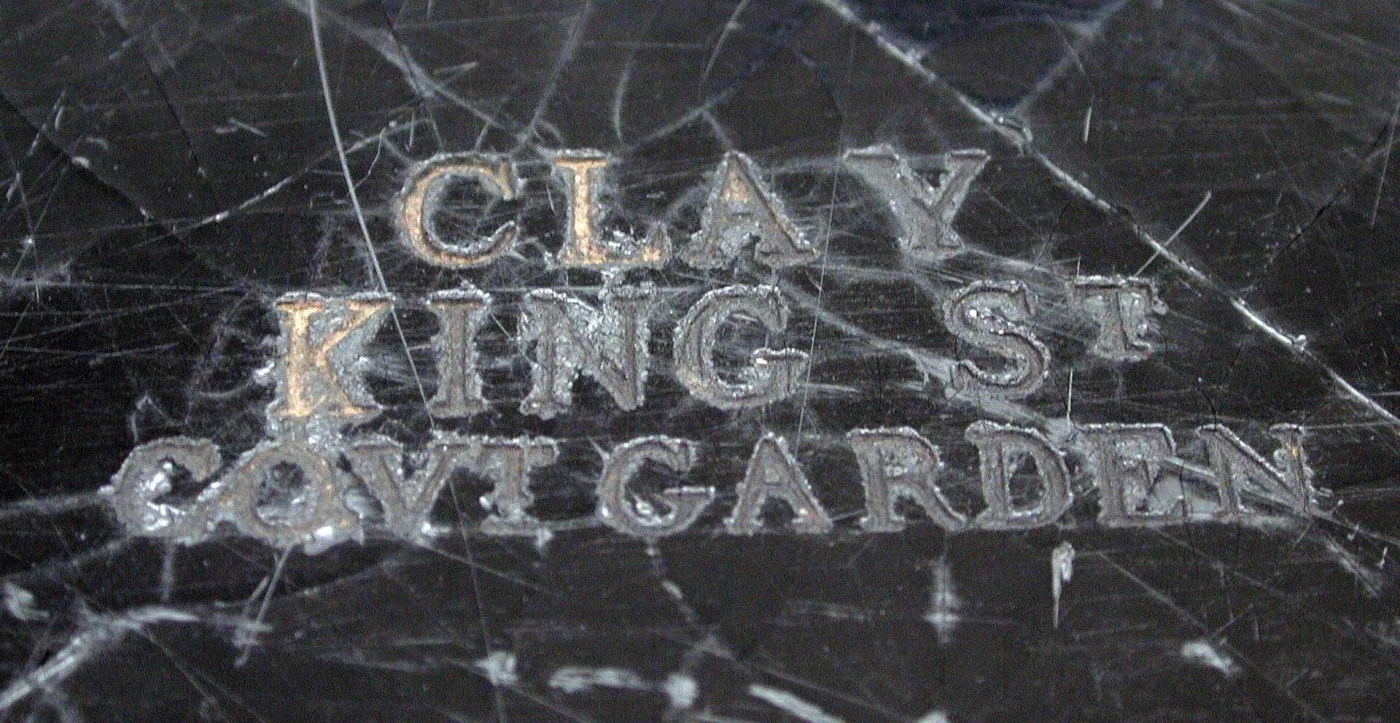 Close view of a dark scratched surface stamped with three lines of small capital letters reading CLAY, KING ST, COVT GARDEN
