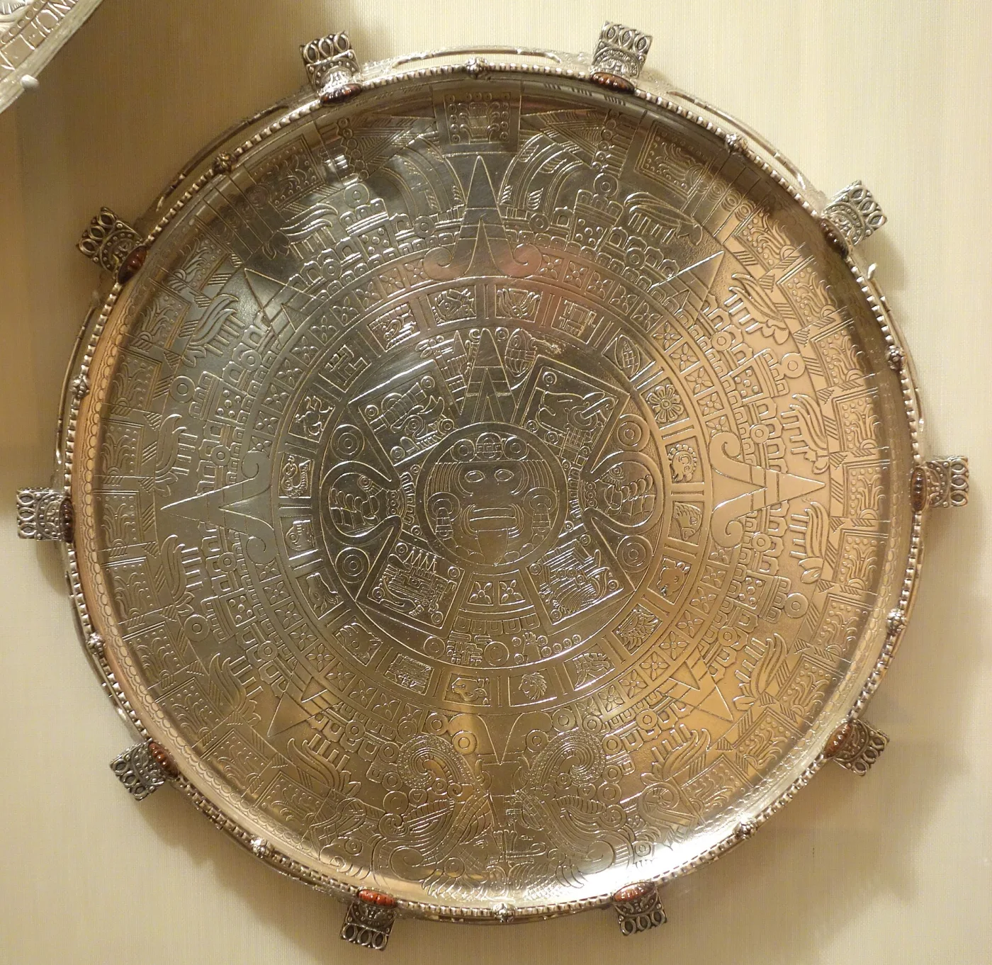 Circular silver tray engraved overall with the concentric bands and central face of the Aztec sun stone, with twelve applied openwork mounts set with polished stones around the rim