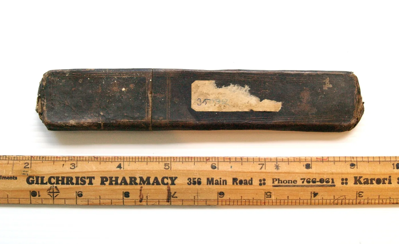 Straight razor with a pale bone handle and a heavily oxidised blade, lying above its black cardboard case, both laid on an advertising ruler