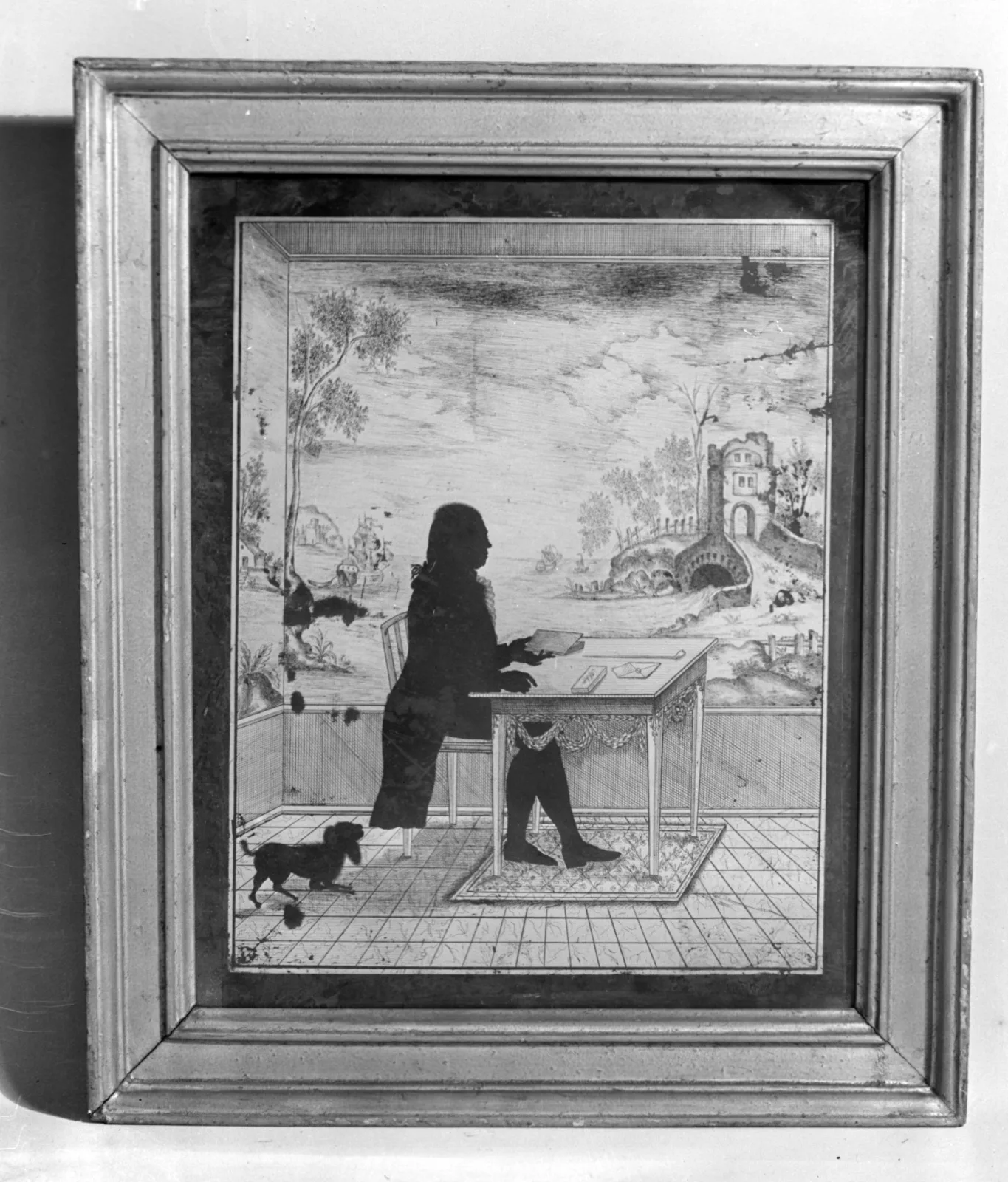 Framed silhouette panel in a moulded gilt frame showing a seated figure at a table in a fully drawn interior with a dog on a tiled floor, the figure solid black and the setting rendered in fine line