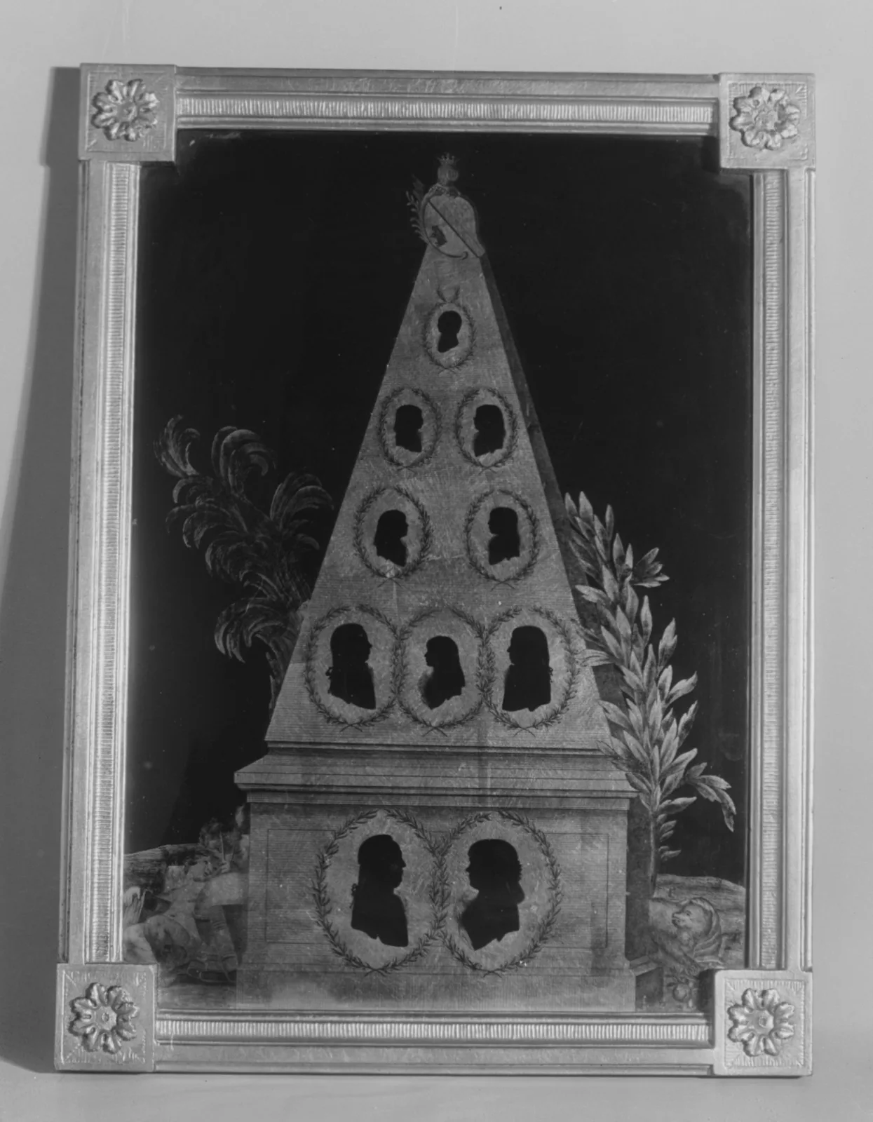 Tall obelisk-shaped mount holding a family group of silhouettes: two large busts side by side at the base with eight smaller oval profiles arranged in rows above them, all under glass in a frame