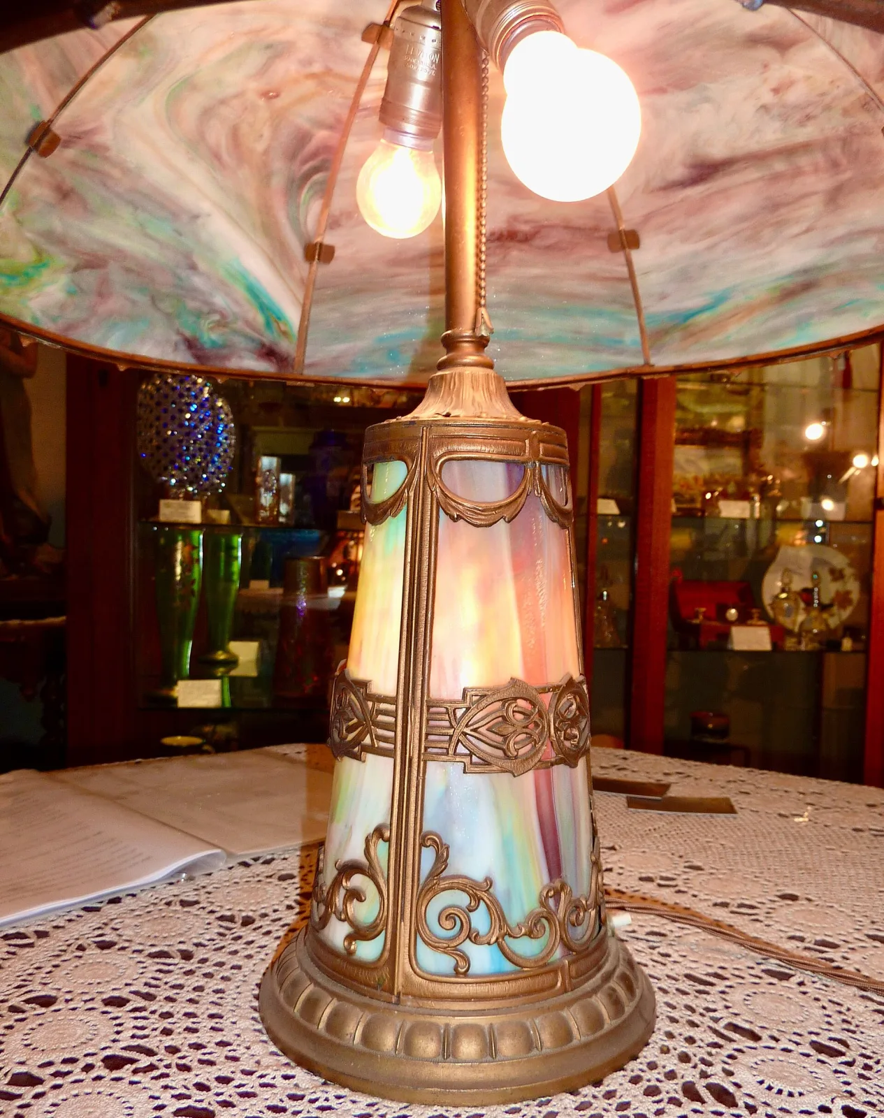 The same lamp lit and seen from below, showing the smooth unpainted inside of the shade with metal clips holding each panel, and a tapering illuminated base of slag glass panels in a metal frame