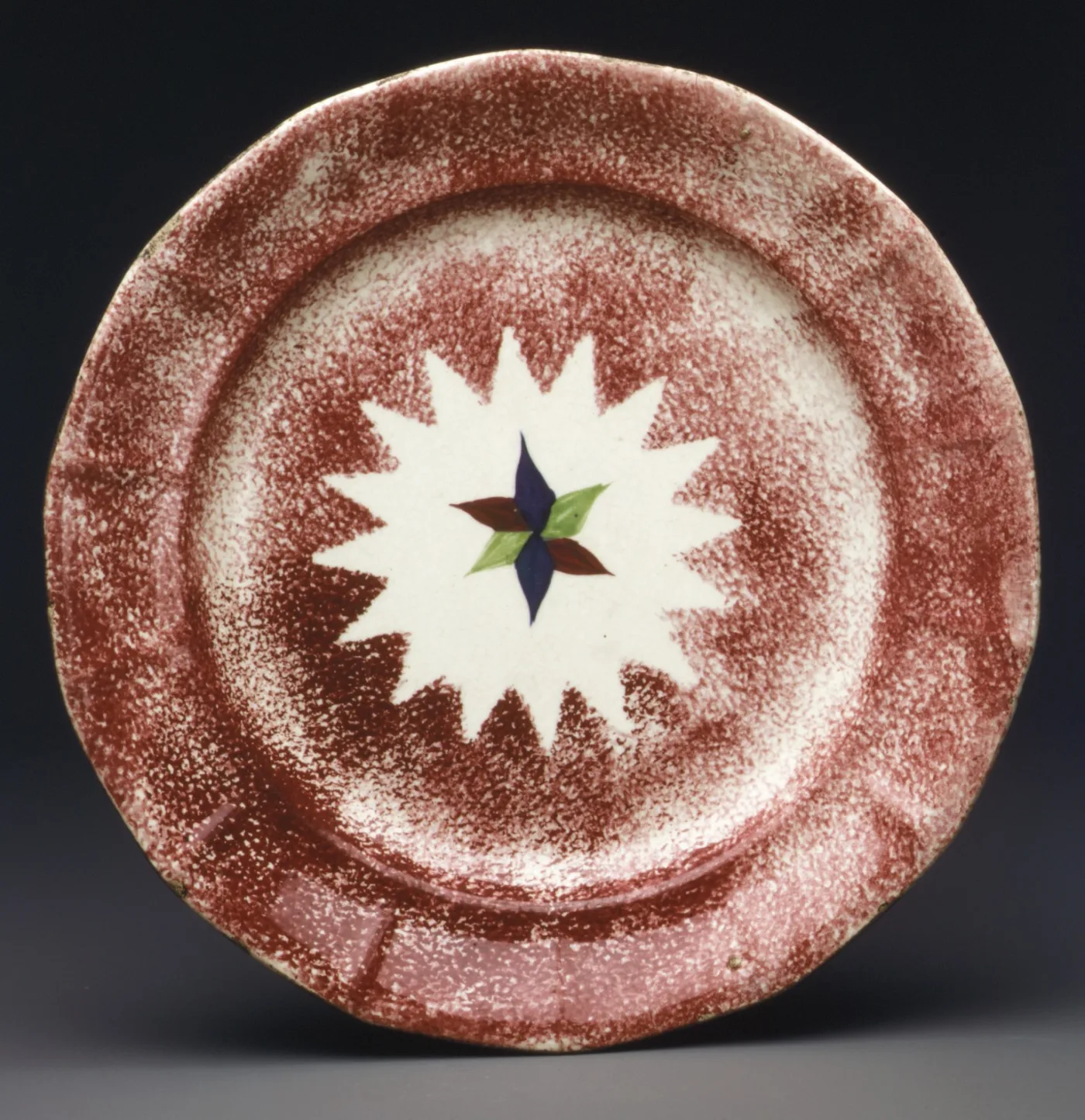 A plate with a dense red sponged border surrounding a reserved white star shape, with a small hand-painted four-petal motif in blue, green and brown at the centre