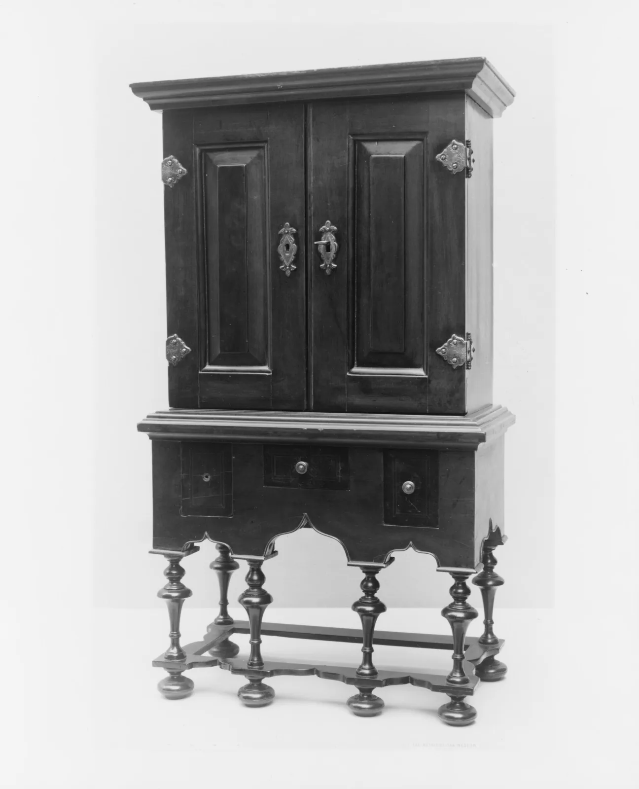 Archival photograph of a small two-door cabinet standing on six turned legs, with a shaped skirt, three drawers below the doors and pierced hinges