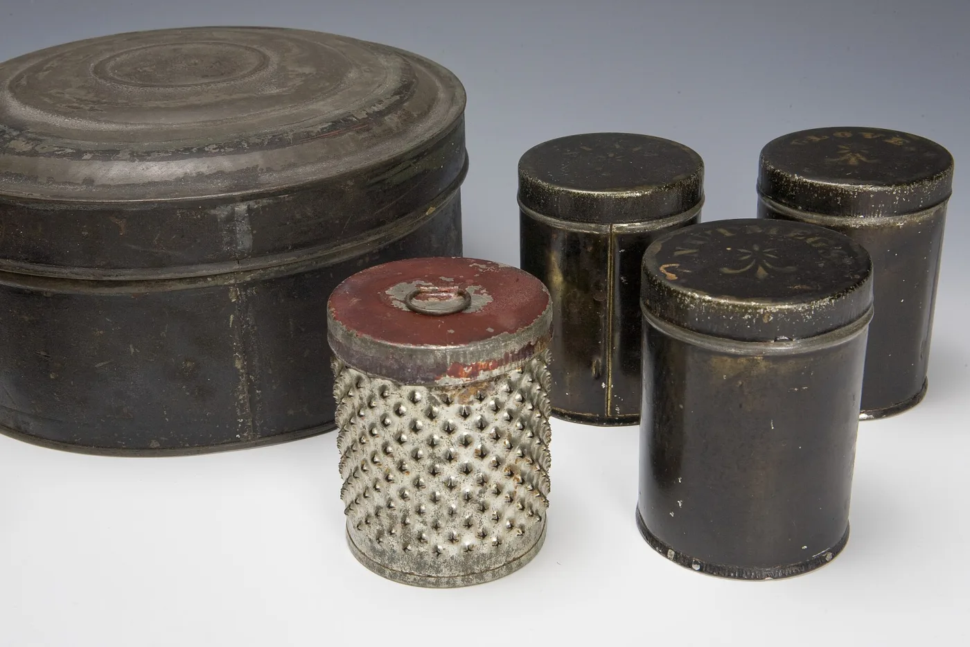 Dark painted tin spice box with its domed lid beside three cylindrical spice canisters and a punched-tin grater cylinder with a red lid