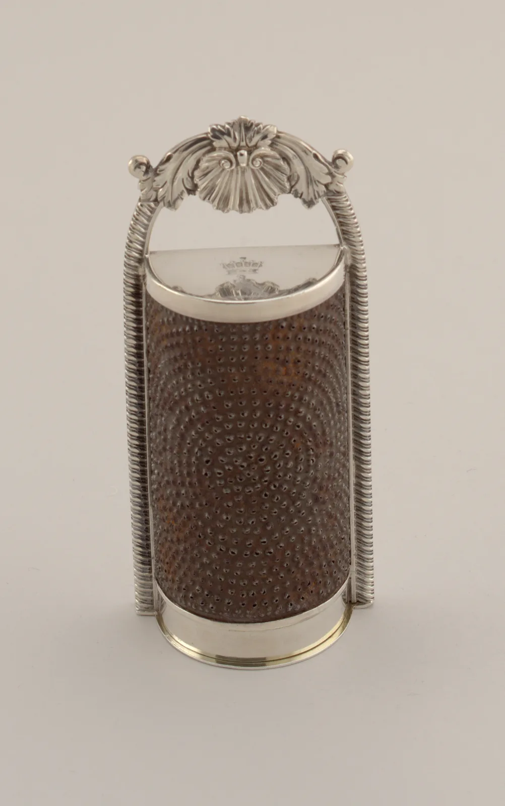 Silver-mounted nutmeg grater with a shell and acanthus crest above two reeded uprights, a brown pierced grating cylinder held between them, and small hallmarks struck on the silver lid