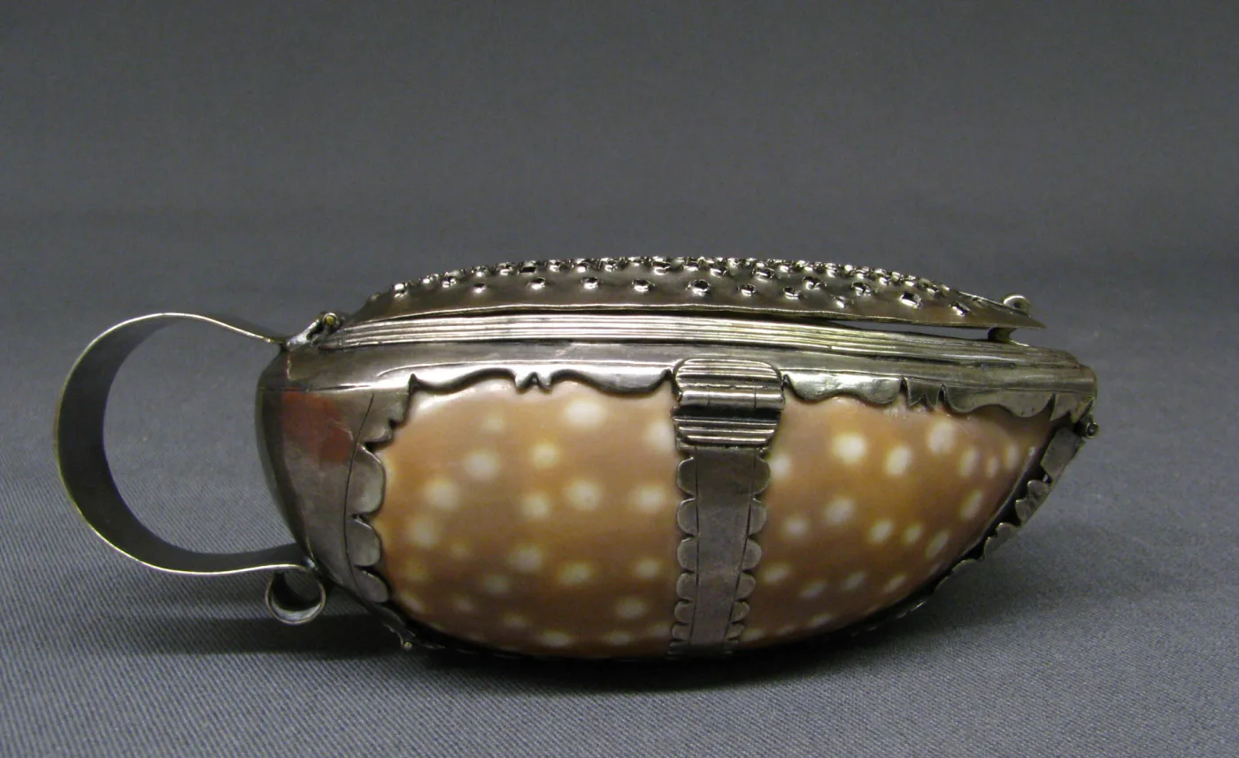 Small oval nutmeg grater made from a spotted sea shell mounted in scalloped silver, with a pierced grating plate hinged into the lid and a looped silver handle