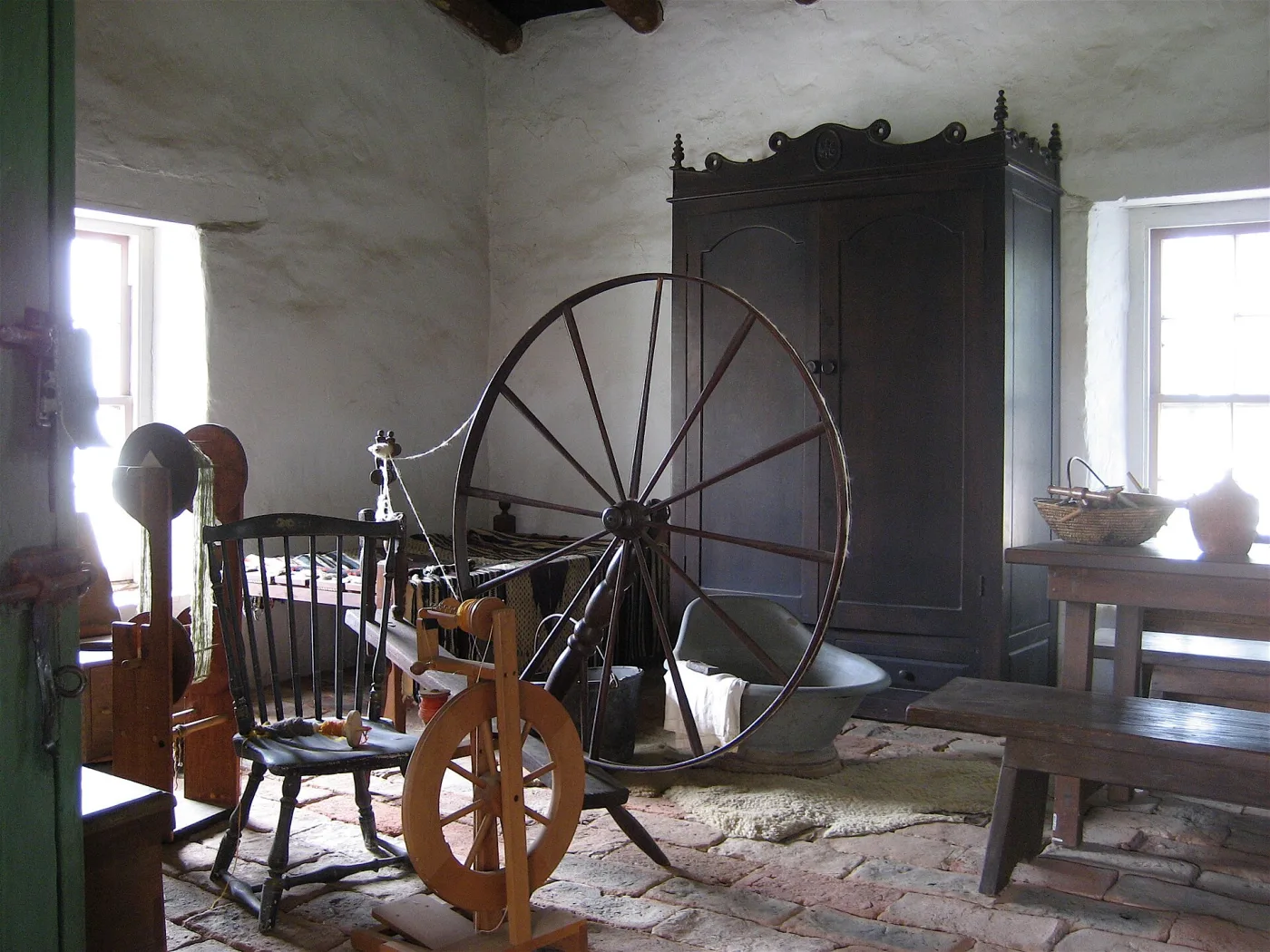 Room with two spinning wheels: a large dark spoked wheel on a raked bench behind, and in front a smaller pale wheel with a treadle, upright posts and a flyer, beside a loom and a swift