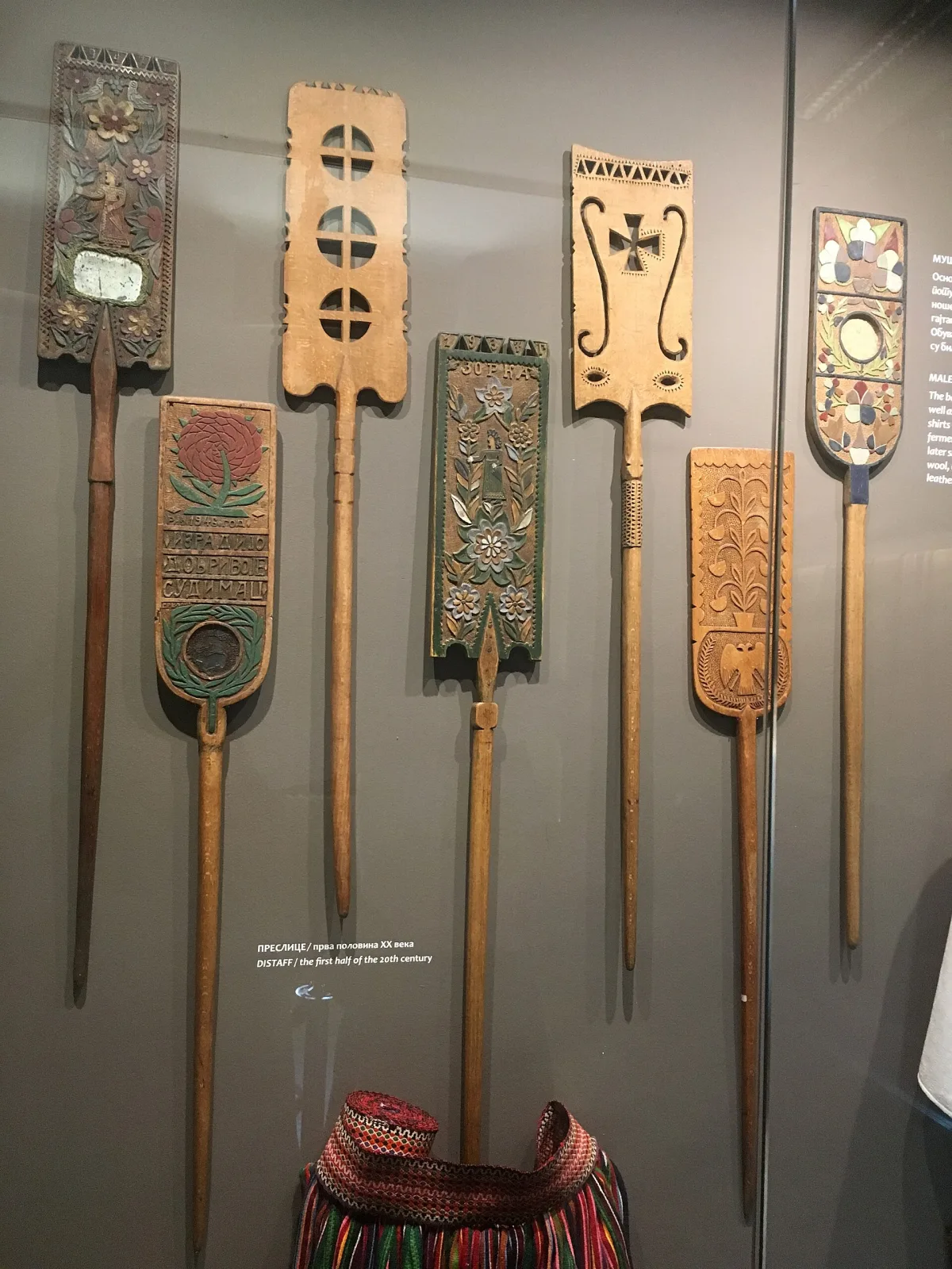 Seven carved wooden distaffs mounted side by side on a museum wall, each a flat decorated blade on a long tapered shaft, variously chip-carved, pierced, painted and inset with a mirror or a coin