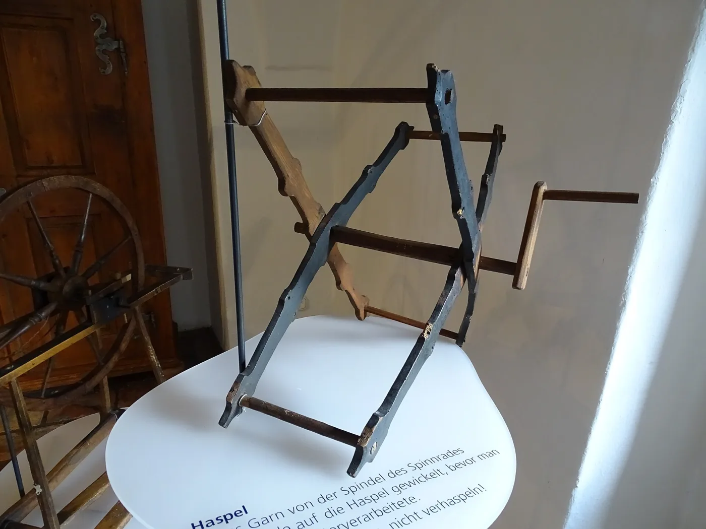 Wooden skein winder with a blue-grey painted cross frame, four brown crossbars and a turned crank handle, displayed on a white museum plinth with a printed label, a spinning wheel behind it