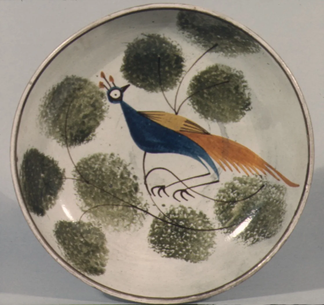 Early nineteenth century saucer painted with a blue and orange peafowl perched on a thin branch, surrounded by rounded blobs of green foliage applied with a sponge