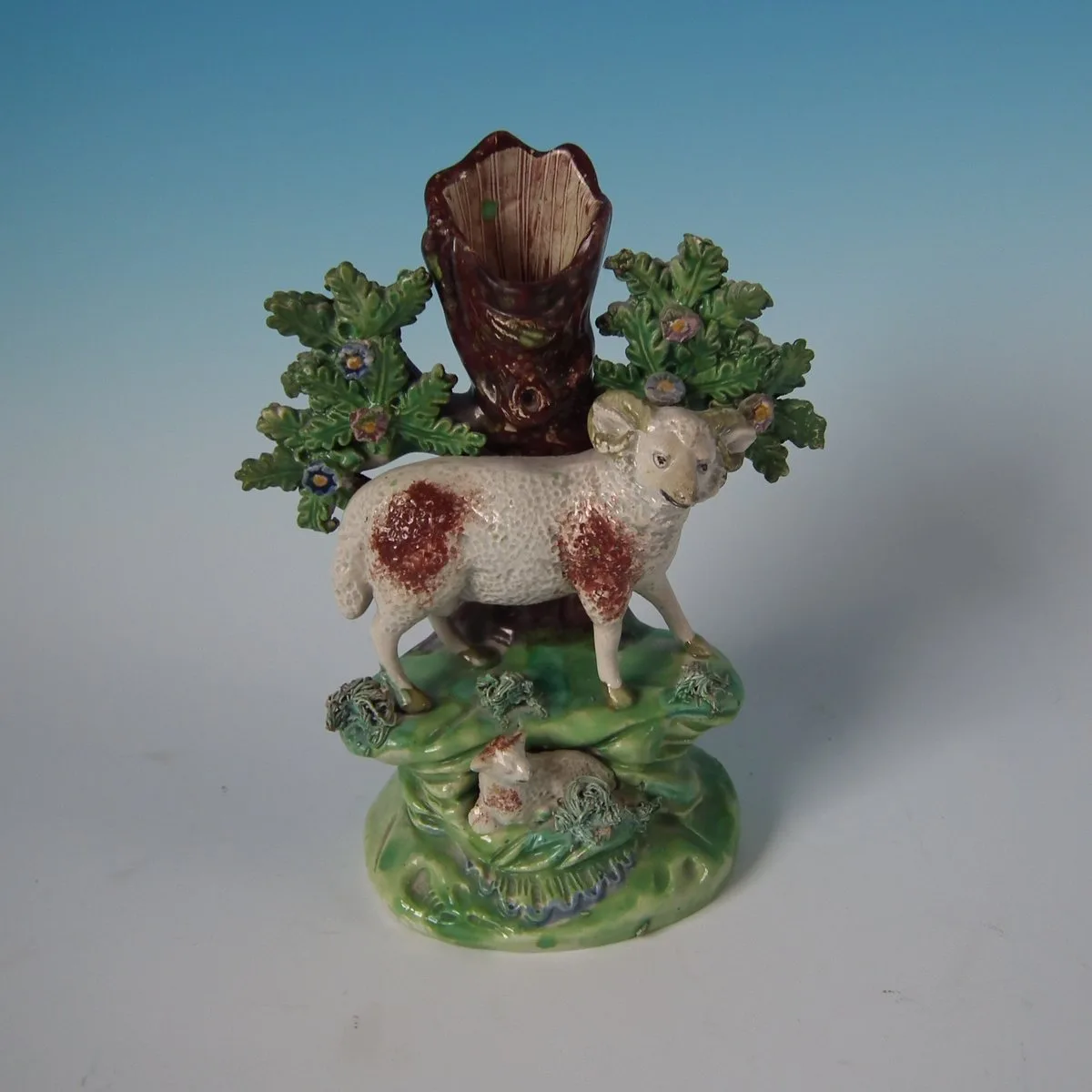 Pottery ram with rust-coloured patches standing before a hollow tree trunk flanked by oak-leaf bocage, a lamb lying on the green base