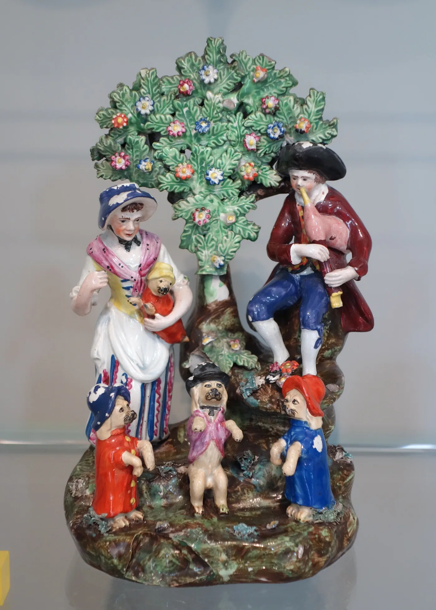 Pottery group of a piper and a woman beneath a leafy tree, with three small dogs in coats and hats standing on the base