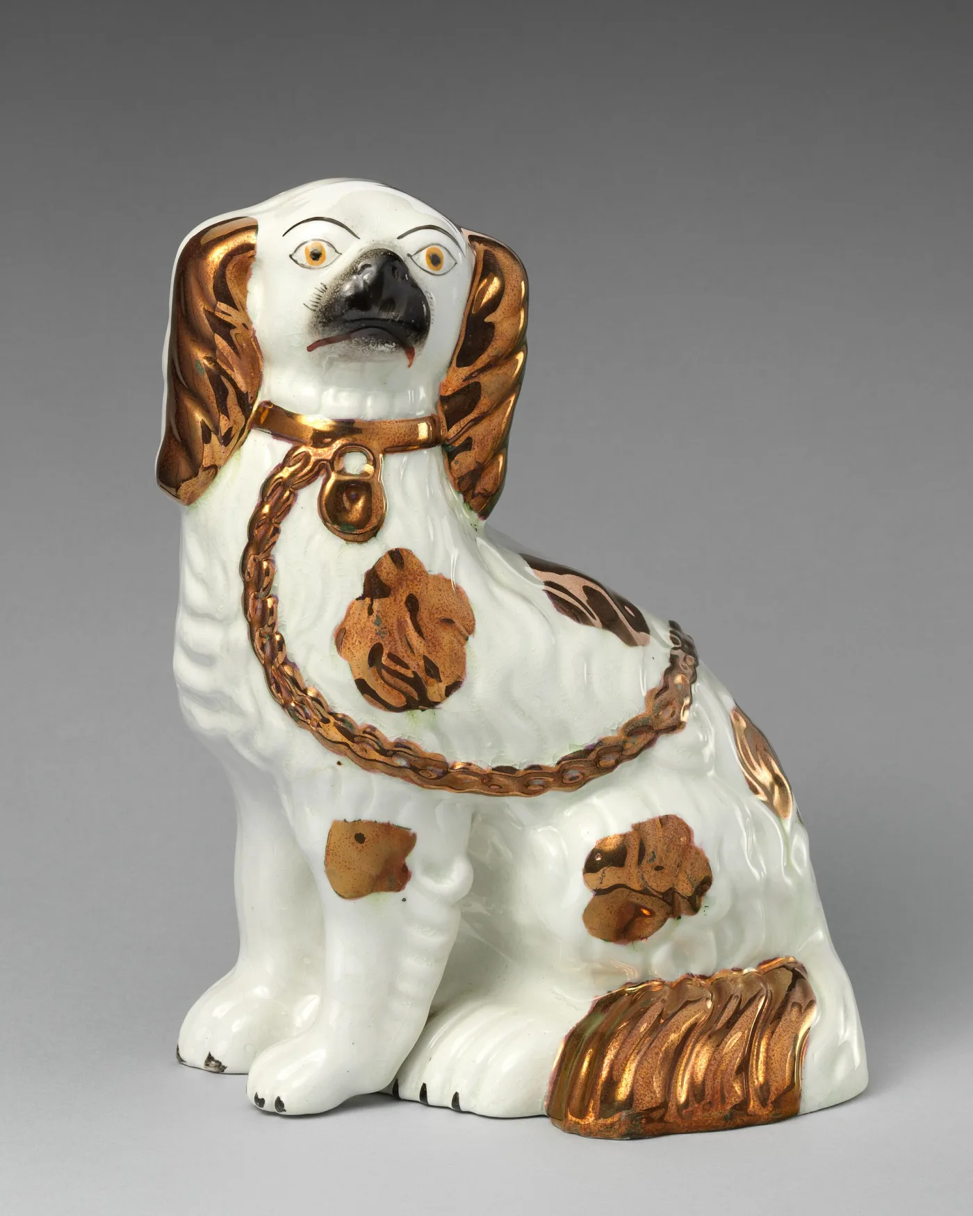 Seated white pottery spaniel with copper-coloured patches, a gilt chain and padlock at its neck, and a black muzzle