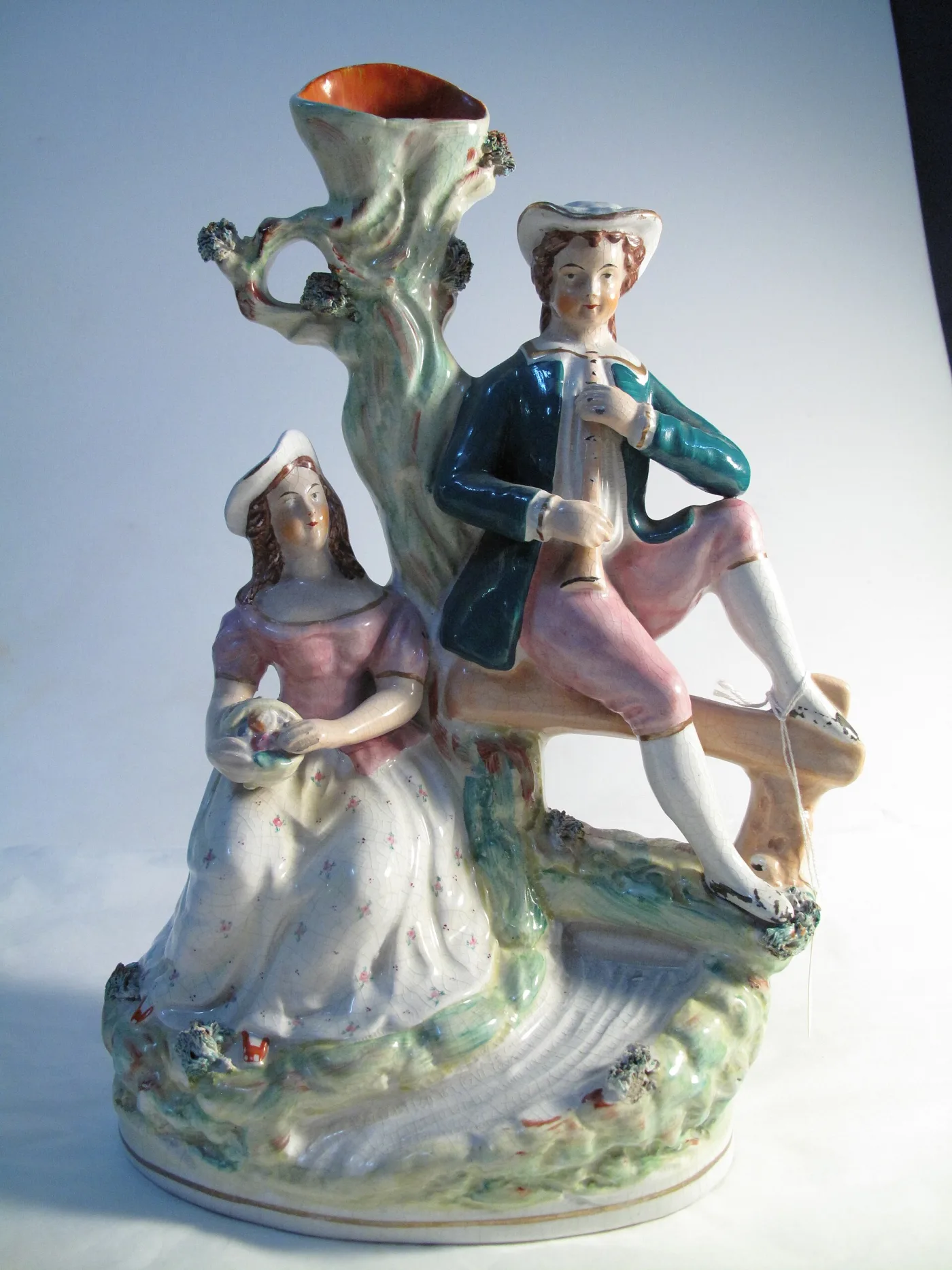 Front of a pottery group: a piper seated on a fence and a woman in a flowered dress beneath a tree trunk hollowed into a vase