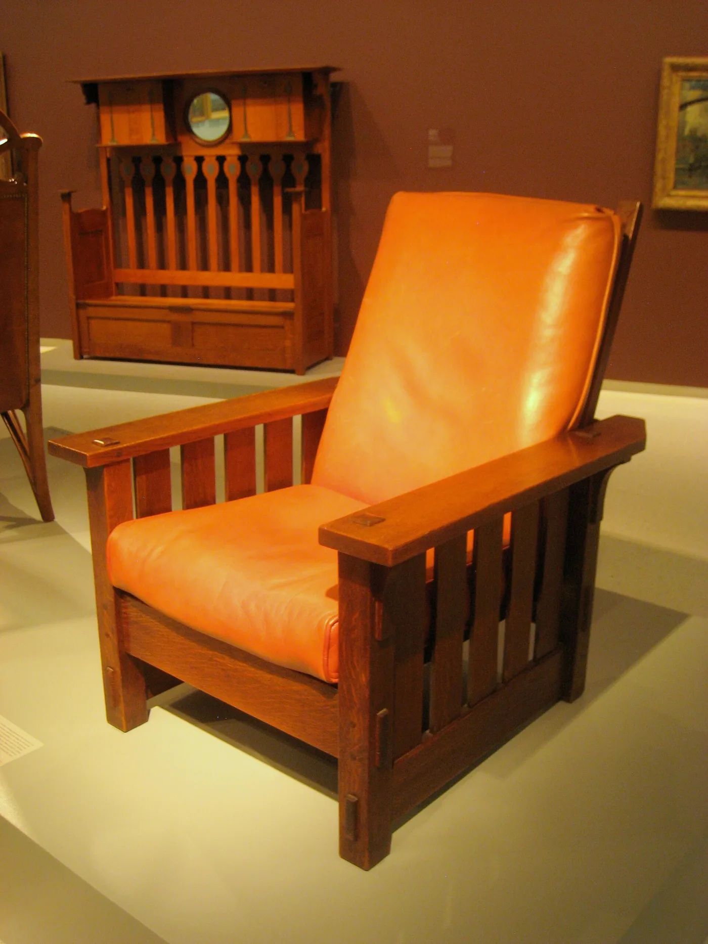 Oak reclining armchair with wide flat arms, vertical slats to the floor and thick tan leather seat and back cushions, displayed in a museum gallery