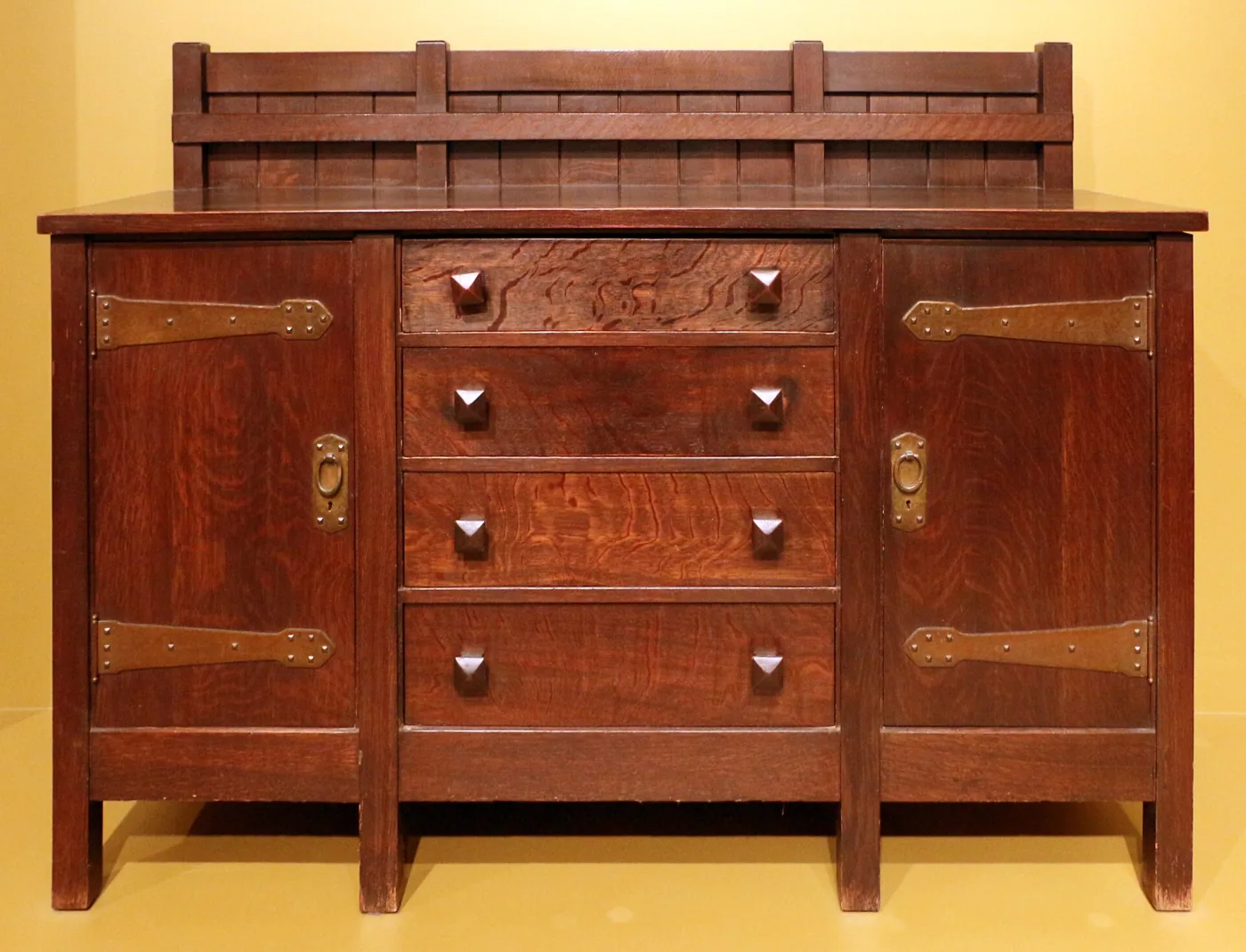 Oak sideboard with a slatted plate rail, two cupboard doors flanking four drawers, long copper strap hinges and pyramidal wooden drawer knobs