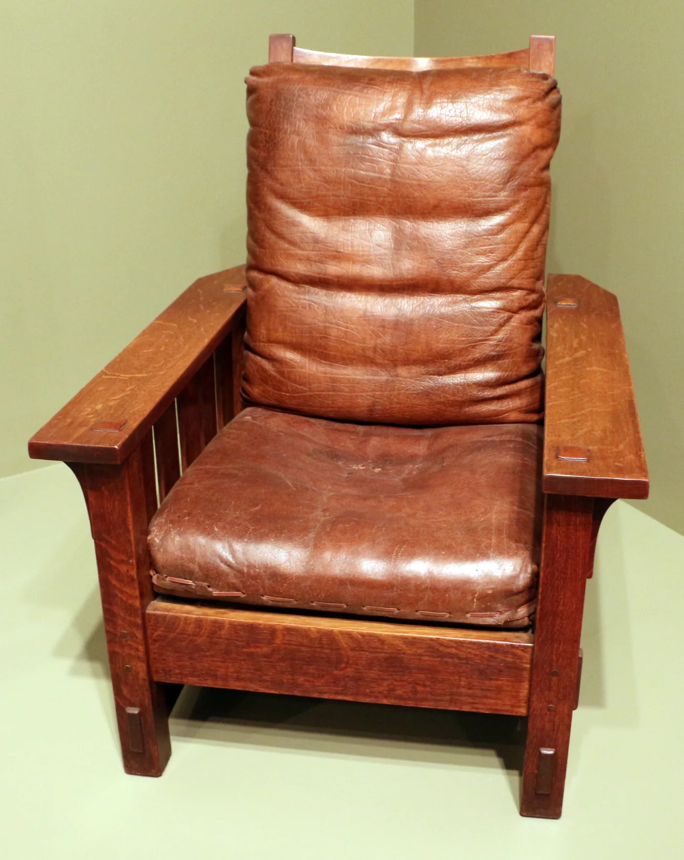 Oak armchair with vertical slats below flat wide arms, worn brown leather seat and back cushions, and square tenon ends showing through the arm tops