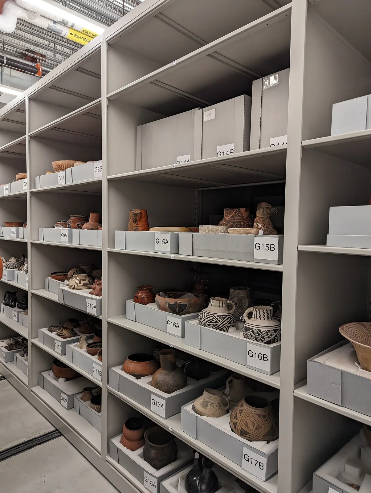 Aisles of grey steel museum shelving holding painted pottery vessels, each standing in its own shallow tray on cut foam, every tray labelled with a location code