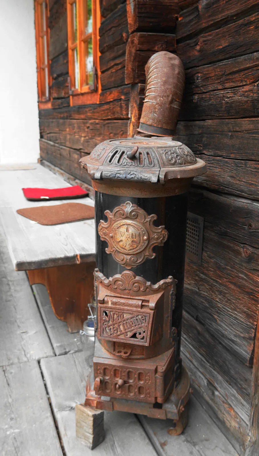 Tall cylindrical cast-iron stove standing against a dark log wall in an alpine farmhouse, with a decorated top section, a rusted flue pipe rising behind it and a cast plate on the black cylindrical body