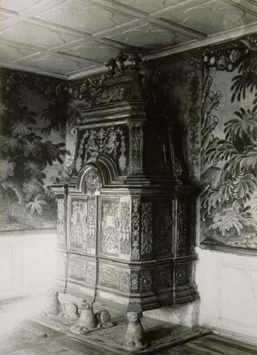 Historic black-and-white photograph of a very large tiered cast-iron stove standing in the corner of a room papered with foliage, its stacked sections covered in relief ornament and supported on cast paw feet