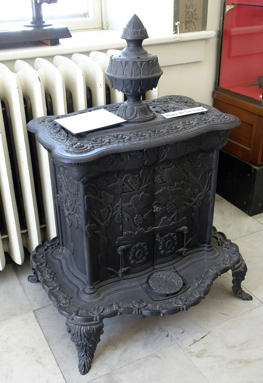 Small black cast-iron parlour stove with a shaped top, a tall urn-shaped finial, low-relief panels on its sides and short cabriole legs, standing in a museum beside a radiator with two label cards on top