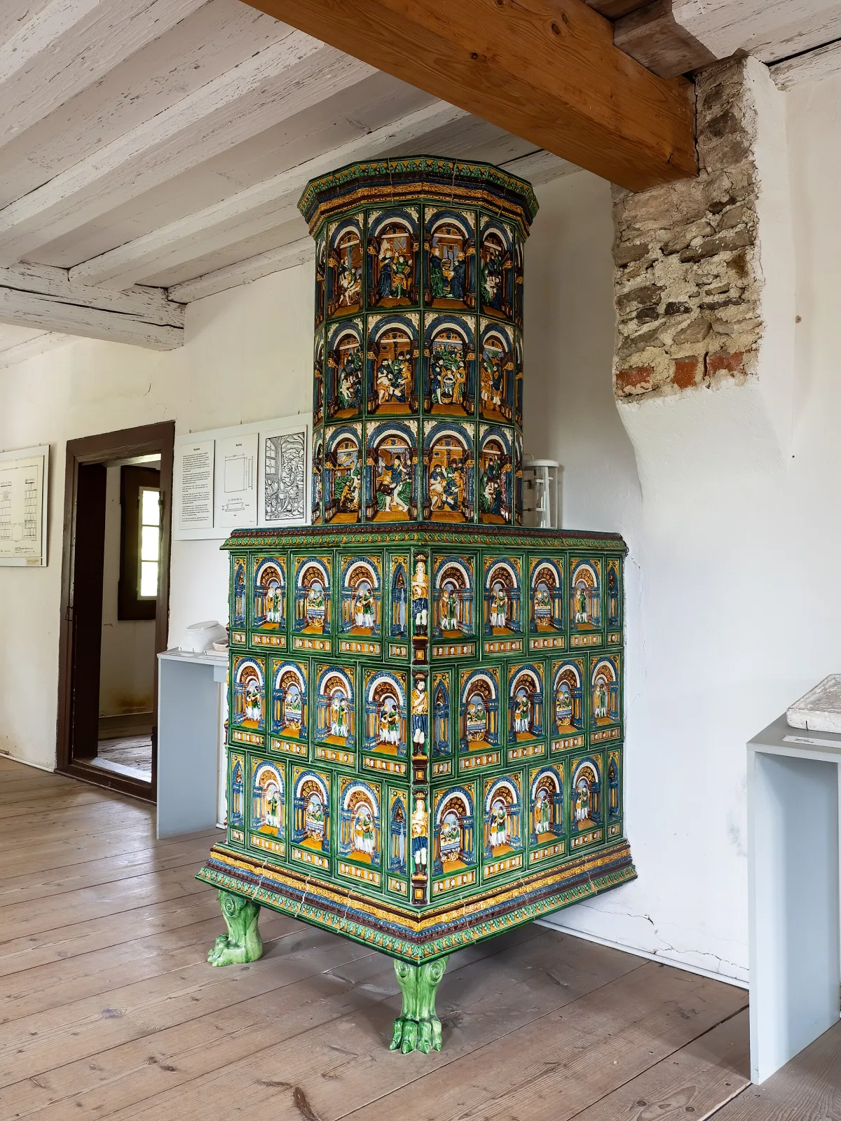 Tall stove of green and polychrome glazed tiles with arcaded panels of figures, standing on green claw feet in a timber-ceilinged museum room