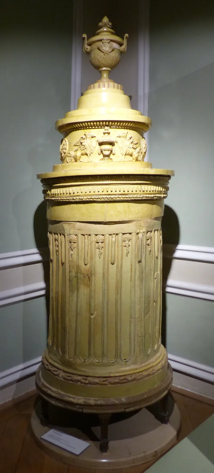 Tall neo-classical tiled stove in pale yellow glazed ceramic, built as a stack of moulded sections with a fluted column-like body and a large lidded urn as its crowning finial