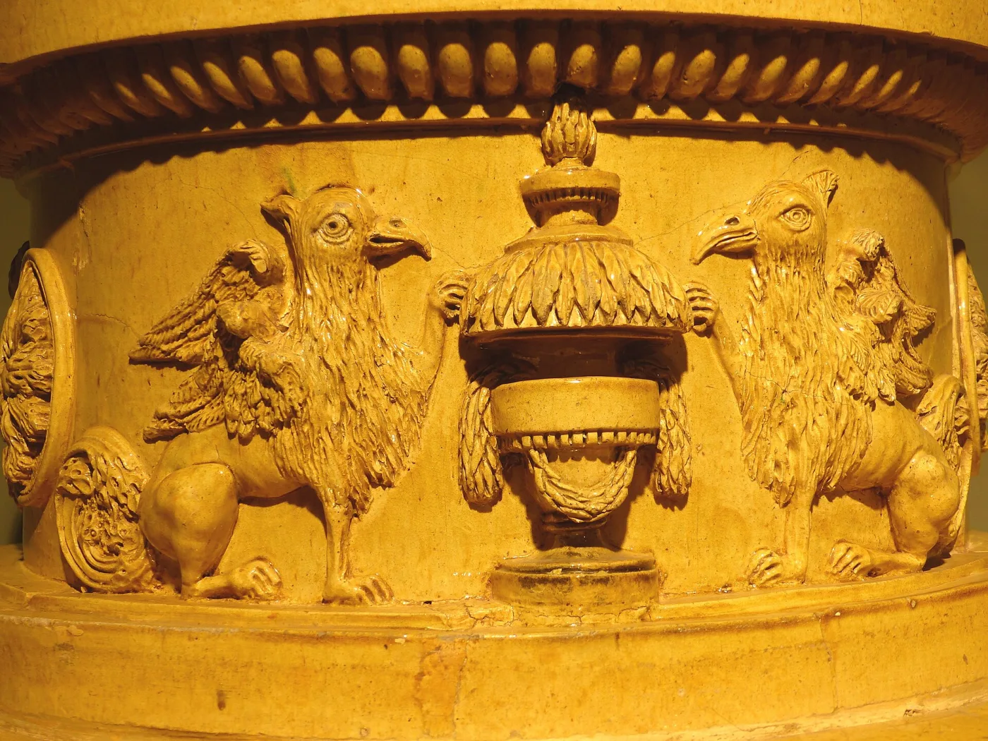 Detail of a yellow glazed stove frieze moulded in relief with two winged griffins facing each other across a draped flaming urn, beneath a band of egg-and-dart moulding