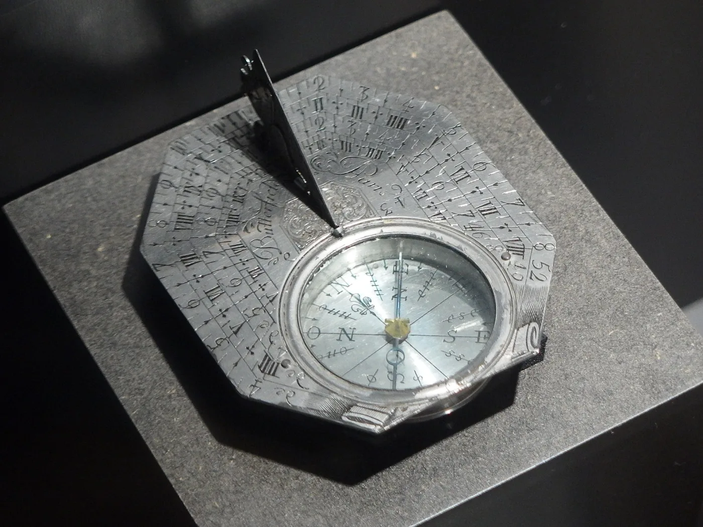 Octagonal silver pocket sundial photographed at an angle in a display case, the plate engraved Butterfield A Paris in script beside the raised bird gnomon