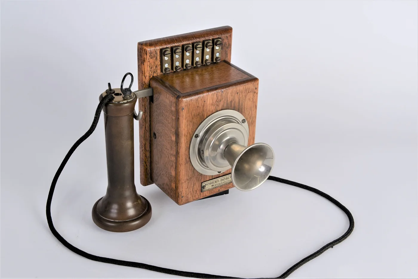 Wall telephone of about 1900 in a polished wooden case, with a crank handle on the right side, two brass bells above a hinged writing shelf, a hook-hung receiver on a cord and a fixed transmitter mouthpiece on a bracket