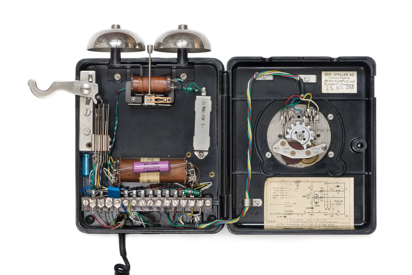 The same wall telephone opened, showing two bell gongs, a row of coils and capacitors, a terminal block with numbered screws, the dial mechanism from behind and a printed service label inside the case