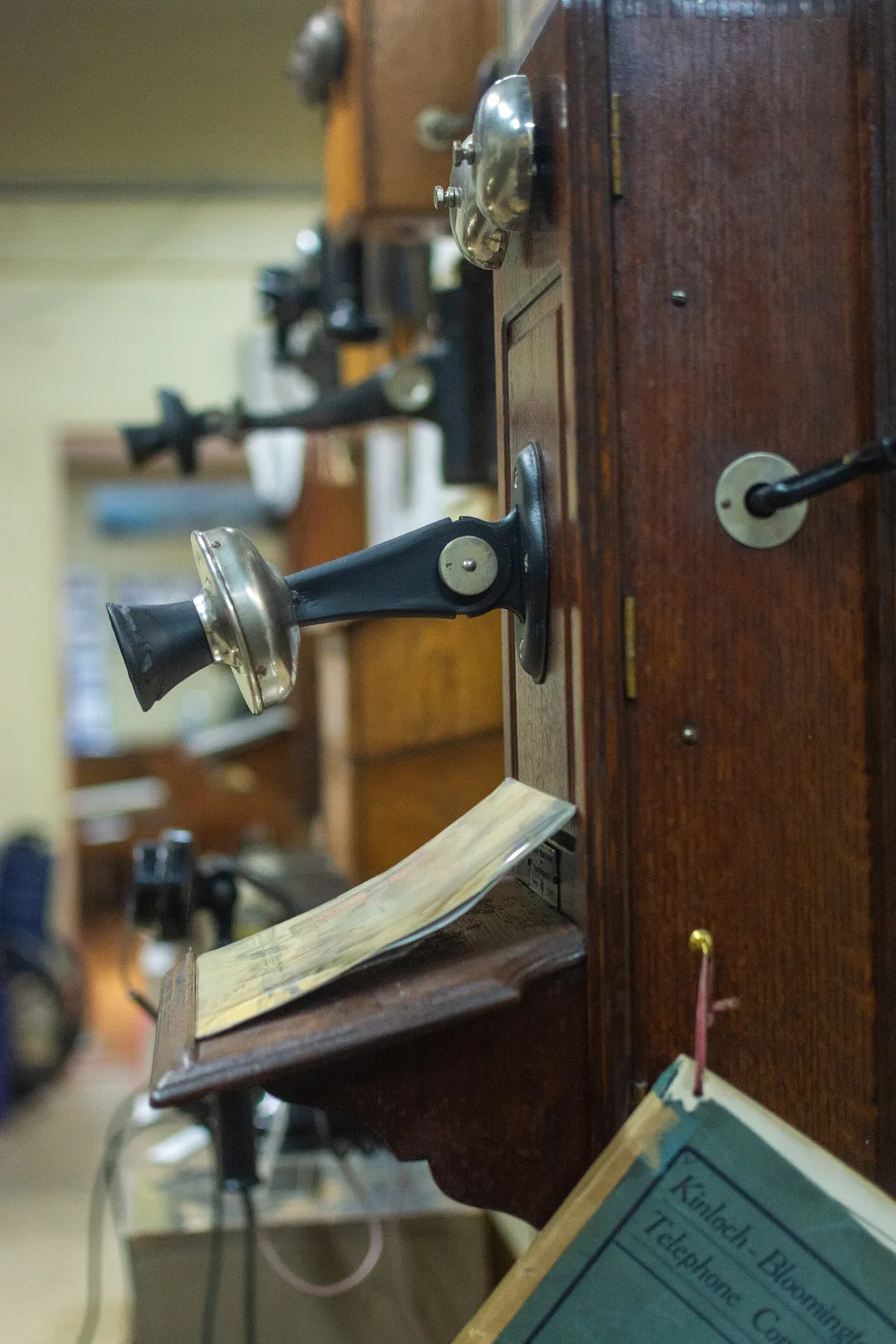 Tall wooden crank wall telephone mounted beside a door in a museum, with a black receiver hanging on its hook, a projecting transmitter arm and a hinged shelf, seen at an angle