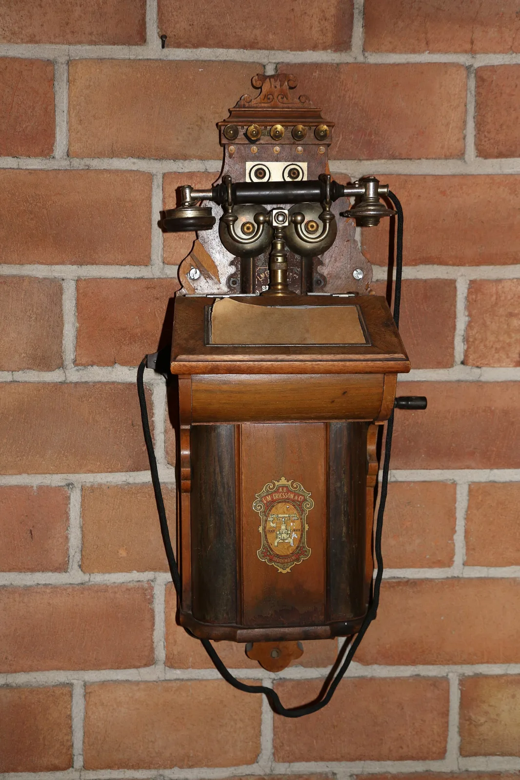 Ornate wall telephone with a polished copper-coloured case, a decorative pierced crest above the bells, a receiver on a curved hook and a crank at the side, mounted on a red brick wall