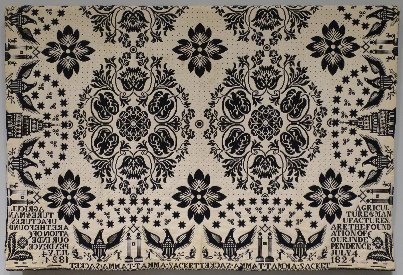 Corner of an 1824 blue and white jacquard coverlet, woven with large stellar flowers, eagles and buildings, and a border of lettering that reads in mirror image