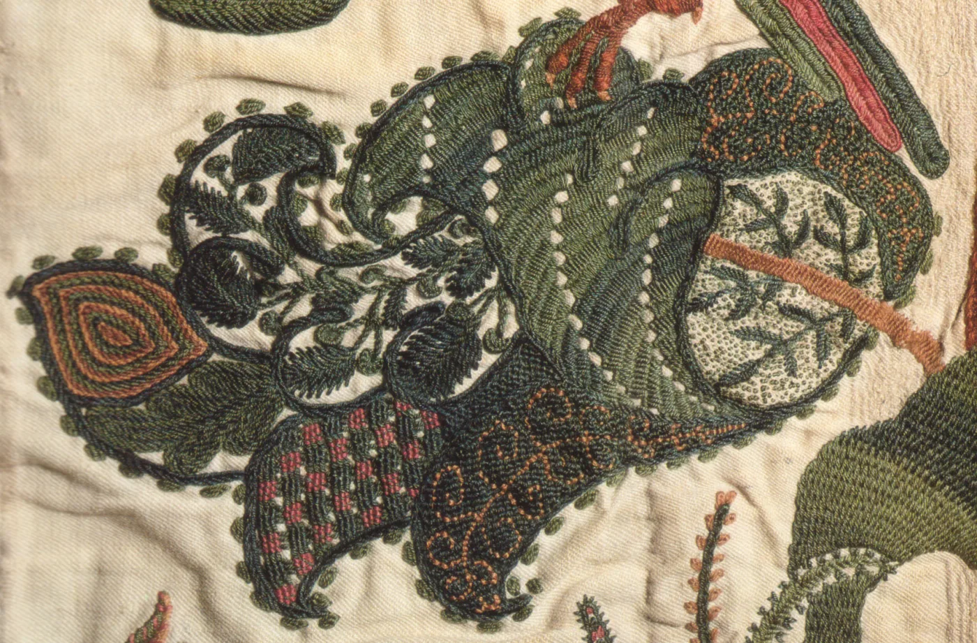 Close detail of crewel embroidery on a linen ground, a large fanciful leaf worked in bands of green, blue and coral wool with visible stitch direction against the plain woven ground beneath