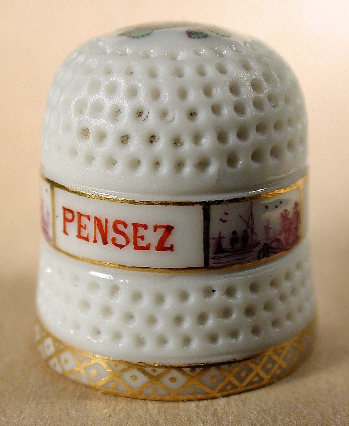 Eighteenth-century Meissen porcelain thimble, its white body covered in moulded dimples, with a red band lettered PENSEZ round the middle above a painted landscape panel and a gilt border at the rim