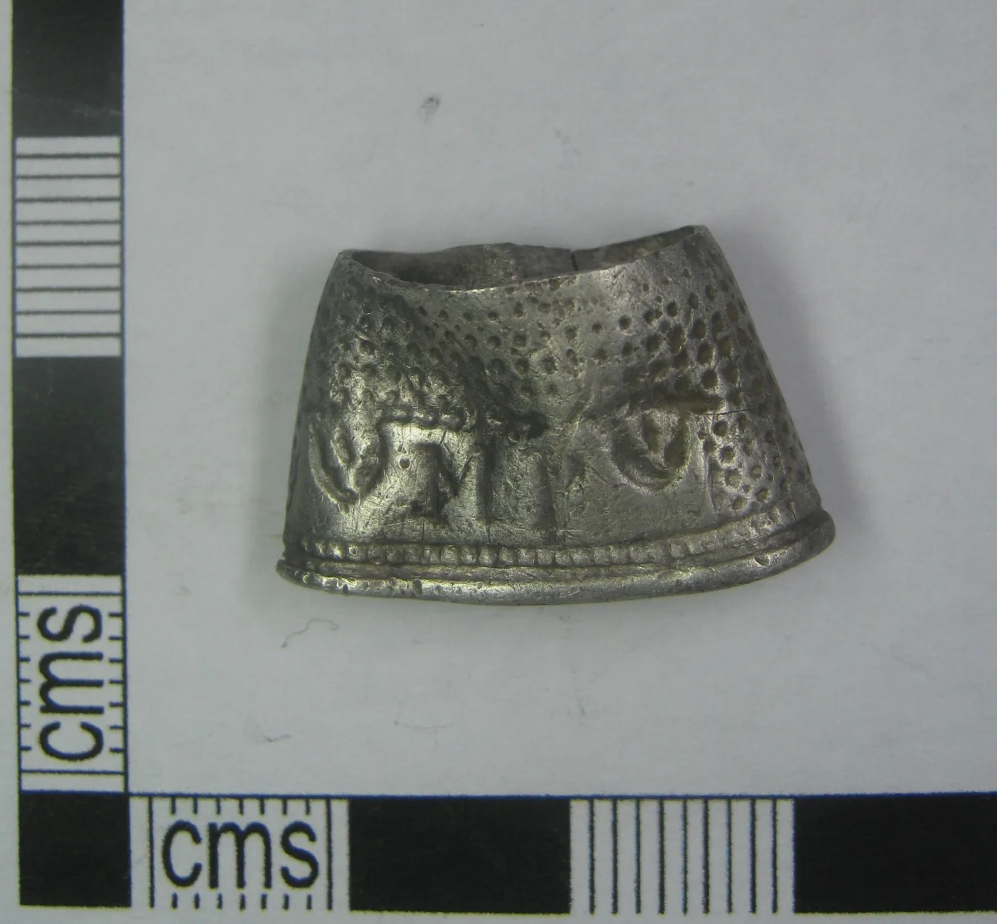 Squat seventeenth-century silver thimble seen from the front against a grey ground with a centimetre scale, its surface covered in dense punched pits and a plain rim below