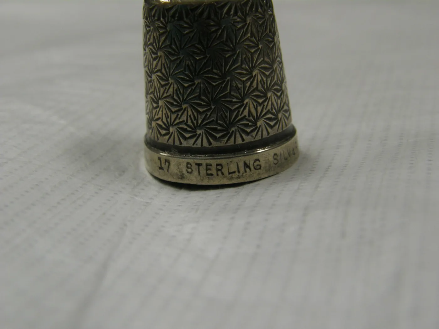 Silver thimble standing on white cloth, its sides patterned with a radiating design and the plain band round its base carrying stamped lettering