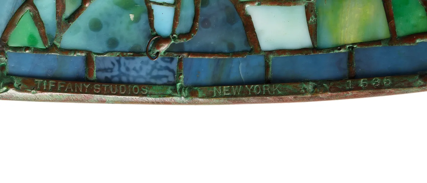 Close view of the lower rim of a leaded glass shade, the bronze band impressed TIFFANY STUDIOS in serif capitals below rows of mottled blue and green glass held in soldered leading