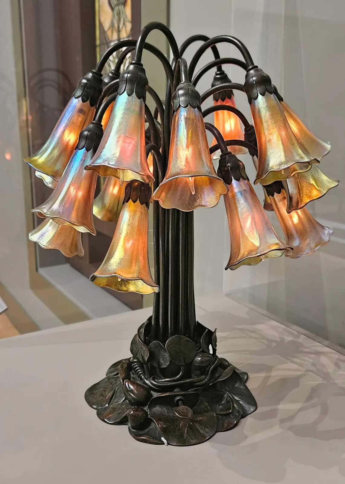 Eighteen-light lily lamp standing on a plinth, a spray of slender bronze stems each carrying a downturned iridescent glass flower shade, lit, against a plain gallery wall