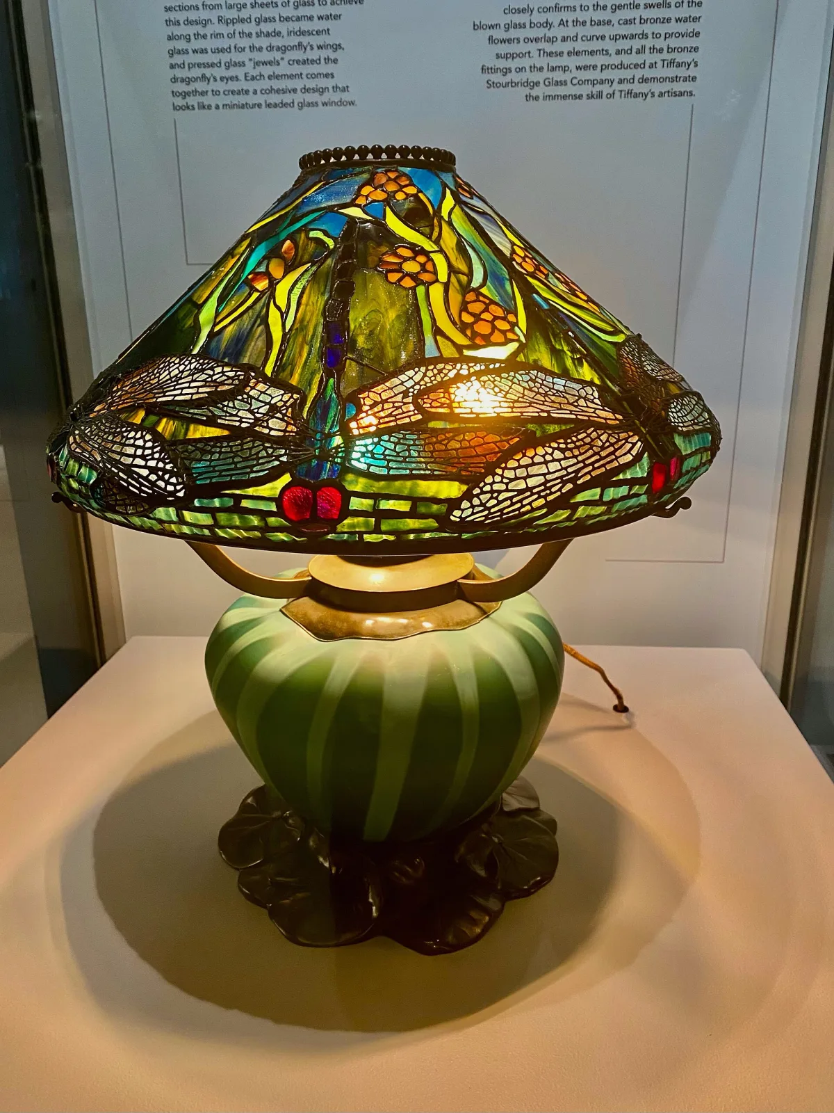 Tiffany reading lamp lit in a museum case, its domed shade patterned with dragonflies and water flowers over a mottled green and amber ground, standing on a swollen green blown-glass base