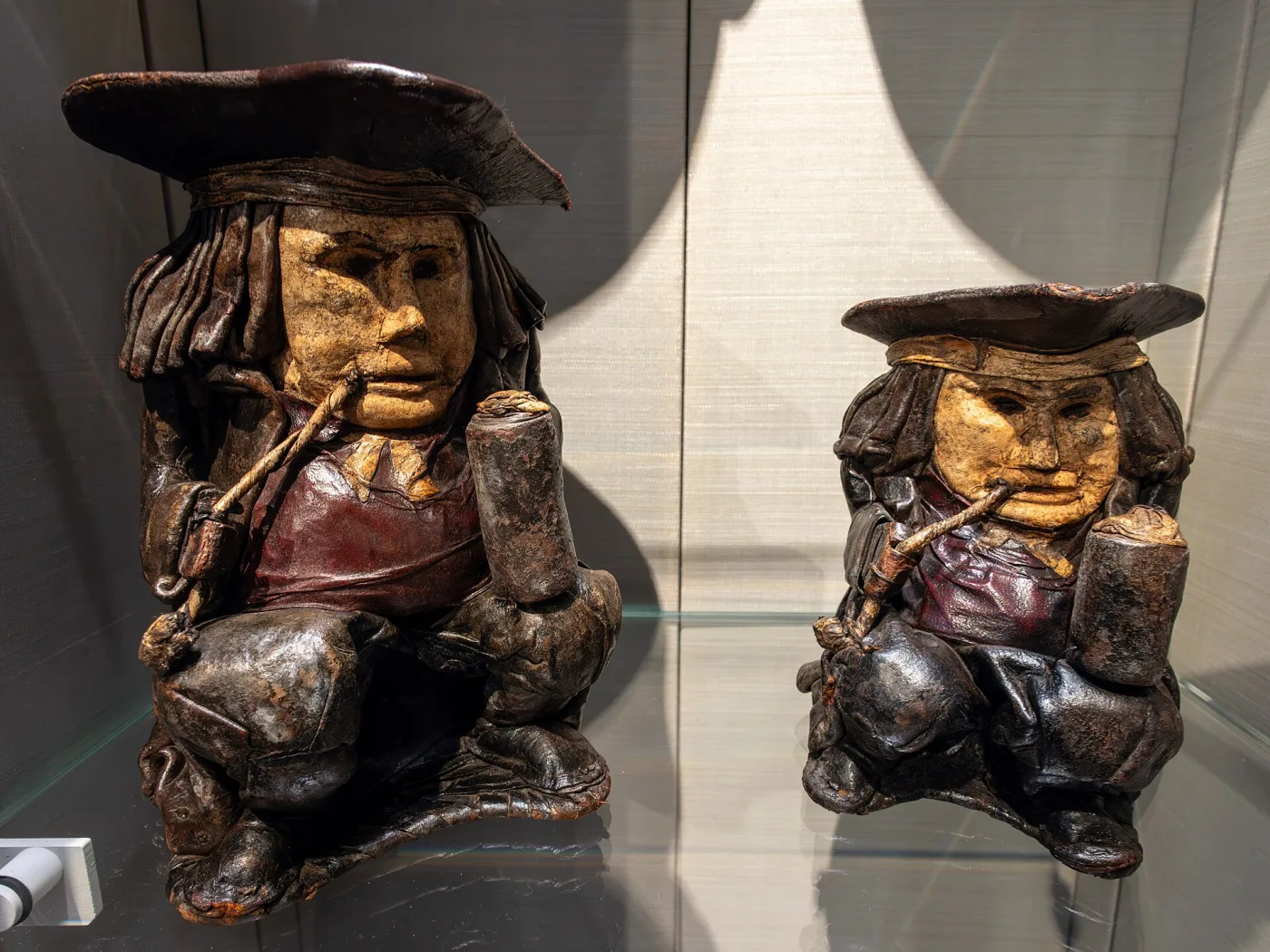 Two dark leather drinking vessels moulded in the form of seated men in tricorn hats, displayed in a lit museum case
