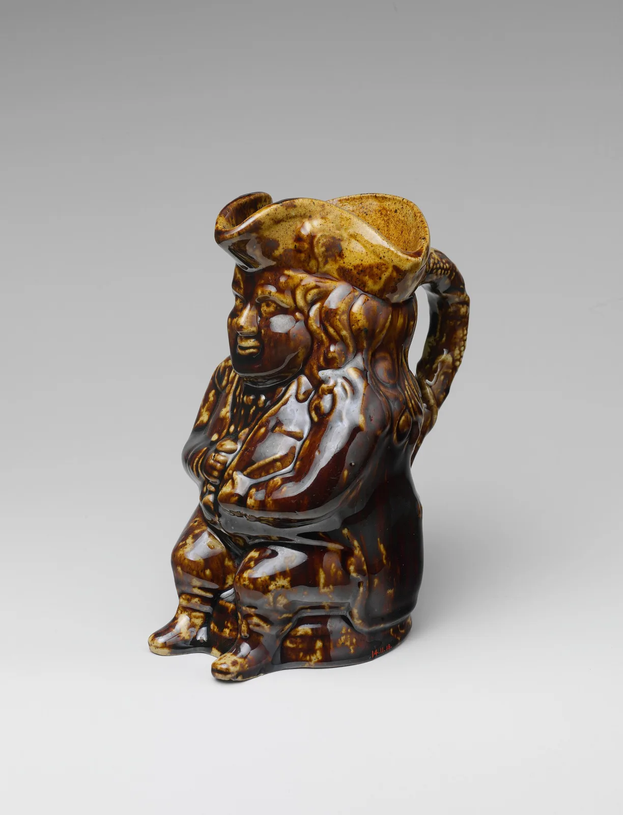 Seated Toby jug of a stout man in a tricorn hat holding a jug on his knee, glazed in streaked and mottled brown, amber and cream